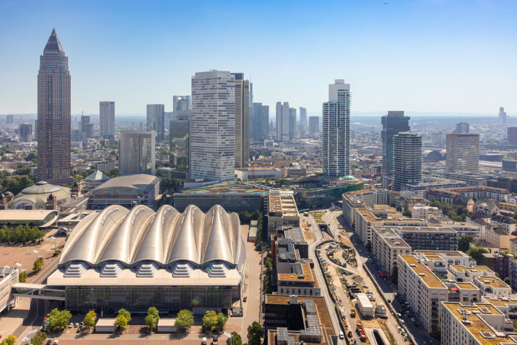Panorama der Frankfurter Skyline mit modernen Hochhäusern, dem Messegelände und umliegenden Stadtvierteln bei klarem, sonnigem Wetter.