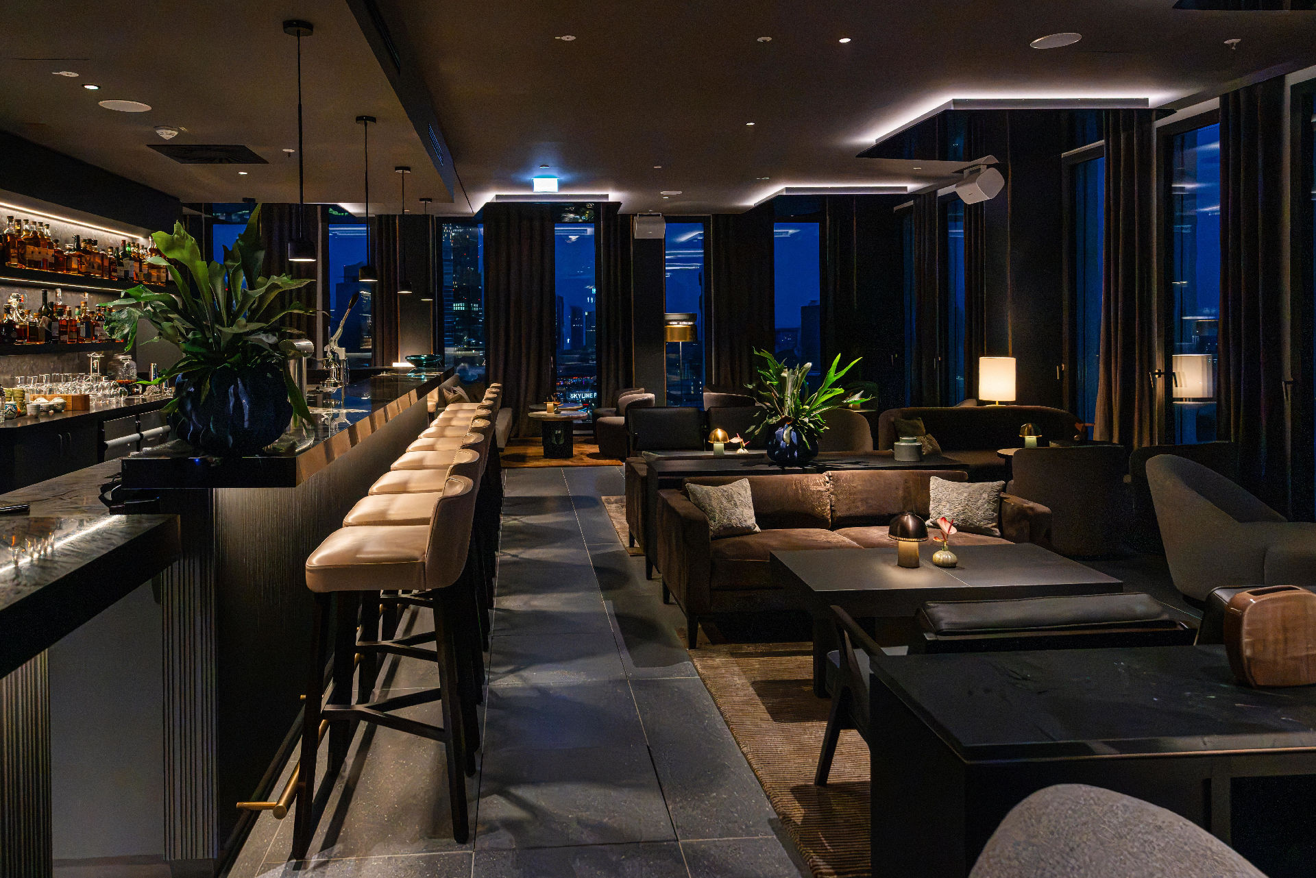 Modern eingerichtete Hotelbar mit langer Theke, hohen Barhockern und gemütlichen Sitzgruppen. Warme Beleuchtung, große Zimmerpflanzen und bodentiefe Fenster mit Blick auf die nächtliche Skyline schaffen eine elegante Lounge-Atmosphäre.