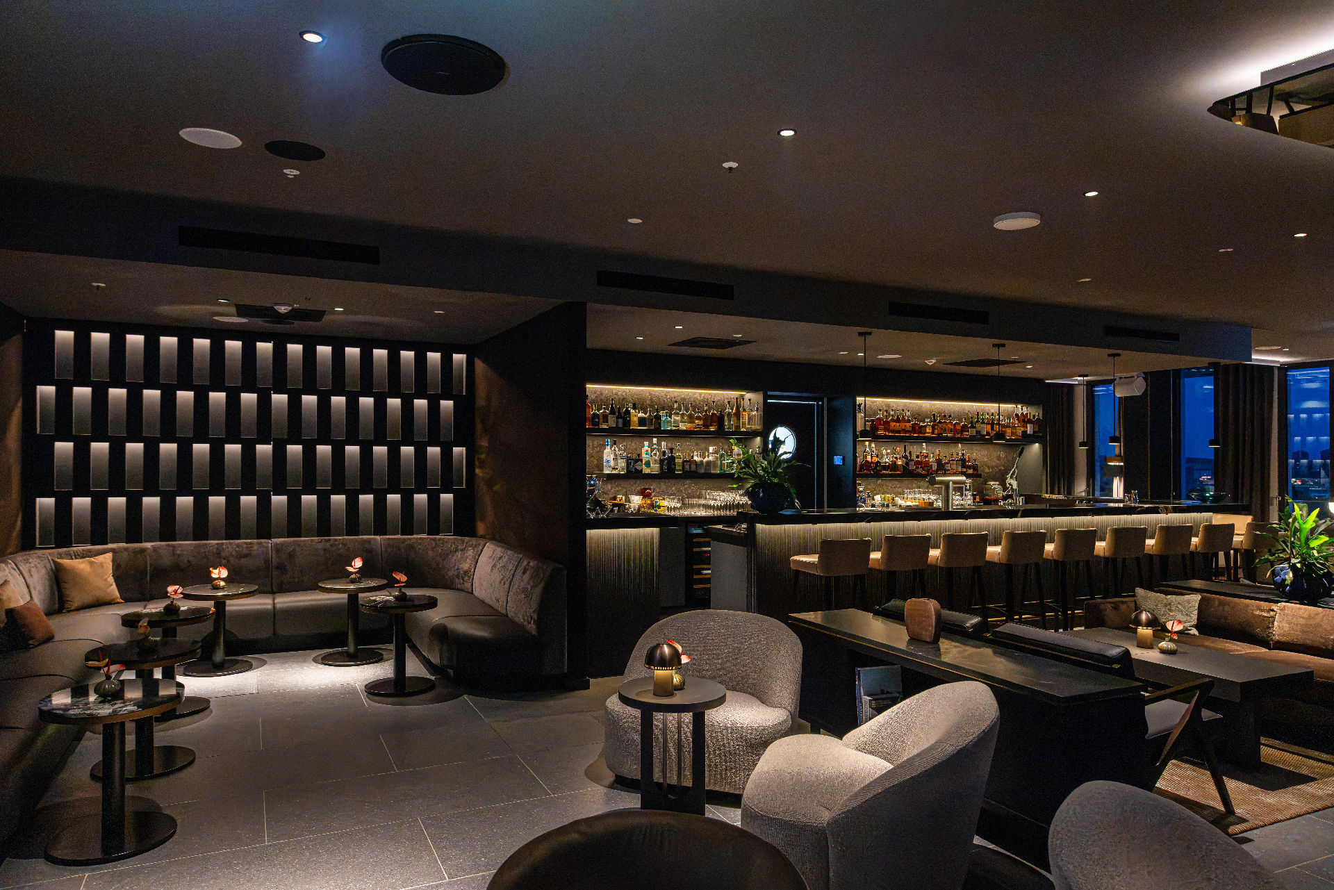 Stilvolle Hotelbar mit langer, beleuchteter Bartheke und hohen Barhockern. Mehrere Sitzbereiche mit Sesseln, Sofas und kleinen Tischen laden zum Verweilen ein. Die dunkle Einrichtung, indirekte Beleuchtung und dekorative Pflanzen schaffen eine elegante Lounge-Atmosphäre mit modernem Design.