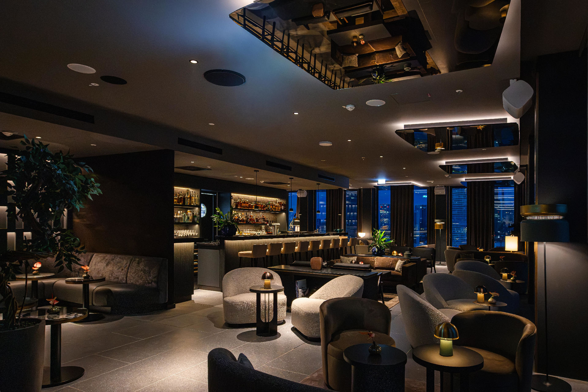 Moderne Hotelbar mit beleuchteter Bartheke, gepolsterten Sesseln und Sofas sowie großen Fenstern mit Blick auf die nächtliche Frankfurter Skyline. Die warme Beleuchtung und das elegante Interieur schaffen eine stilvolle Lounge-Atmosphäre.